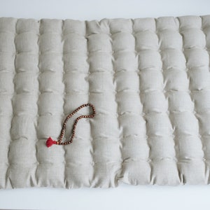 May include: A light beige meditation cushion with a quilted design. A wooden beaded necklace with a red tassel is lying on top of the cushion.