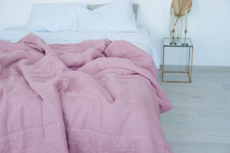 May include: A bed with a light pink linen duvet cover and white sheets. The duvet is draped over the bed, showing the texture of the fabric. A small side table with gold legs and a glass top is visible.