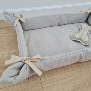 Puede incluir: Una cama para perros de lino gris claro con una almohada con forma de hueso en el interior. La cama tiene un diseño sencillo con un borde fruncido y un lazo atado en la parte superior.