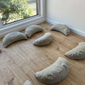 May include: A set of eight crescent-shaped meditation cushions in a light gray fabric. The cushions are arranged in a circle on a light wood floor.