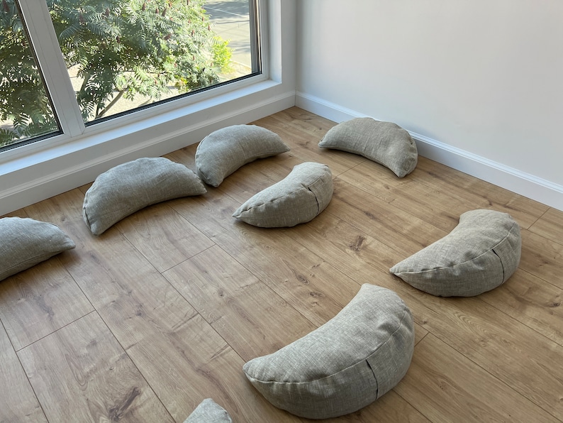 May include: A set of eight crescent-shaped meditation cushions in a light gray fabric. The cushions are arranged in a circle on a light wood floor.