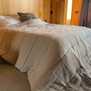 Könnte beinhalten: Ein Bett mit einer beige gesteppten Bettdecke und einem weißen Kissen. Ein flauschiges, strukturiertes Kissen liegt auf dem Bett. Das Bett befindet sich in einem Raum mit Holzwänden und einem grauen Boden. Der Raum hat ein modernes, minimalistisches Design.
