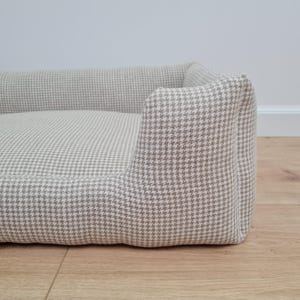 May include: A beige and white houndstooth dog bed with a raised edge. The bed is made of a soft, comfortable fabric and is ideal for small to medium-sized dogs.