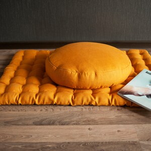May include: A mustard yellow meditation cushion and mat set. The cushion is round and the mat is rectangular with a quilted design. The mat is folded in half and the cushion is sitting on top of it. A book titled "Anticipation - Immunity" is lying on the mat.