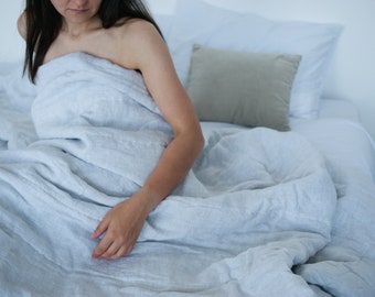 Könnte beinhalten: Ein hellgrauer Bettbezug aus Leinen, der über einem Bett liegt. Der Stoff hat eine sichtbare Webstruktur und ein weiches, knittriges Aussehen. Die Bettwäsche wird mit einem einfarbigen, hellgrauen quadratischen Kissen und einem weißen Kissenbezug kombiniert.