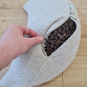 May include: A gray crescent-shaped buckwheat pillow with a zipper opening. The pillow is made of hemp fabric and is filled with buckwheat hulls. The pillow is being held open by a hand, revealing the buckwheat hulls inside.