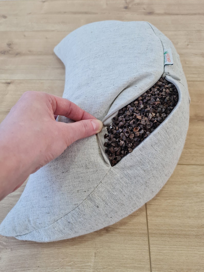 May include: A gray crescent-shaped buckwheat pillow with a zipper opening. The pillow is made of hemp fabric and is filled with buckwheat hulls. The pillow is being held open by a hand, revealing the buckwheat hulls inside.