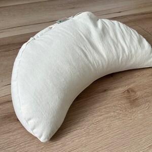 May include: A crescent-shaped meditation cushion in an off-white, textured fabric. The pillow features a curved design with a small fabric tag attached to the top seam, showcasing a visible zipper closure along the edge of the cushion.
