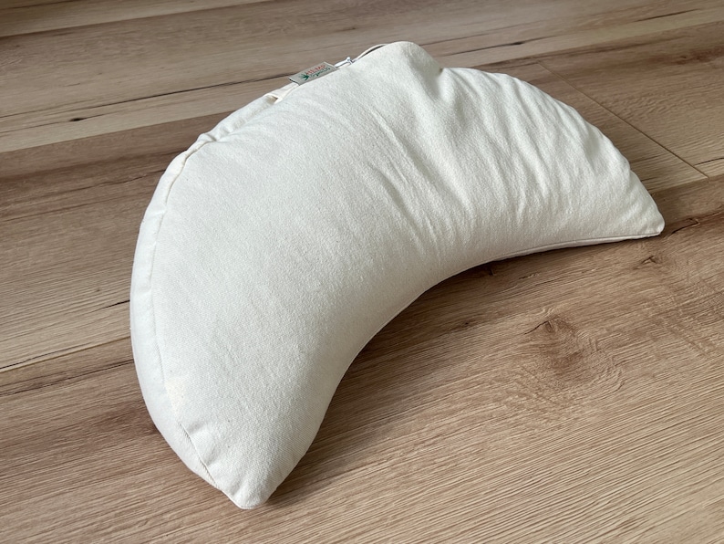 May include: A crescent-shaped meditation cushion in an off-white, textured fabric. The pillow features a curved design with a small fabric tag attached to the top seam, showcasing a visible zipper closure along the edge of the cushion.