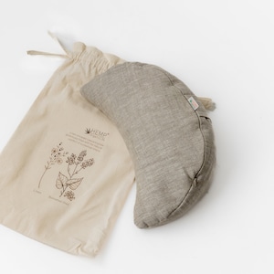 May include: A gray hemp meditation cushion in the shape of a crescent moon, with a hemp drawstring bag. The bag has a printed design of herbs and the text "HEMP ORGANICS".