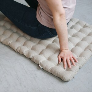 May include: A light beige linen floor cushion with a quilted design. The cushion is rectangular and has a zipper on one side.