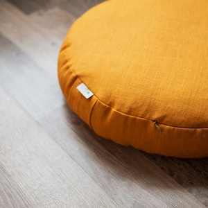 May include: A round, mustard yellow meditation cushion with a zipper closure. The cushion is made of a soft, textured fabric.