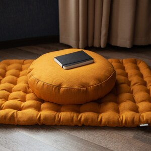May include: A round, mustard yellow meditation cushion with a black notebook on top, sitting on a mustard yellow, quilted meditation mat.