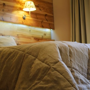 May include: A close-up of a beige quilted duvet on a bed with two pillows. The bed is in a bedroom with a wooden headboard and a lamp on the wall.
