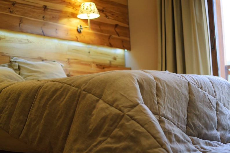 May include: A close-up of a beige quilted duvet on a bed with two pillows. The bed is in a bedroom with a wooden headboard and a lamp on the wall.