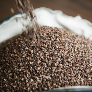 May include: A close-up of a white fabric bag filled with brown buckwheat groats. The groats are being poured into the bag from above.