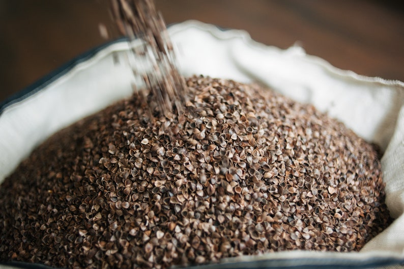 May include: A close-up of a white fabric bag filled with brown buckwheat groats. The groats are being poured into the bag from above.