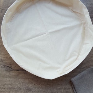 May include: A white cotton round bread proofing basket with a brown linen cloth in the background.
