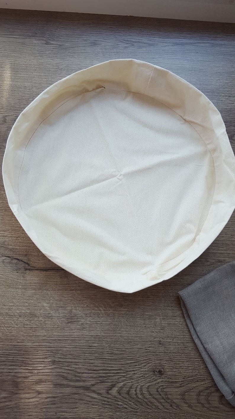 May include: A white cotton round bread proofing basket with a brown linen cloth in the background.