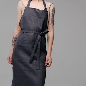 May include: A dark grey linen apron with a single pocket and a tie belt. The apron is being worn by a person with tattoos on their arms.