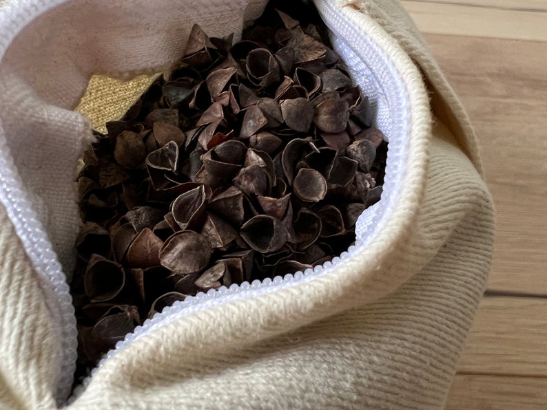May include: A white fabric bag with a zipper closure filled with buckwheat hulls. The bag is lying on a wooden surface.