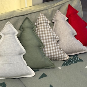 May include: Five tree-shaped decorative pillows in various colors and patterns. The pillows are arranged on a gray sofa and include white, green, plaid, light gray, and red. The pillows are a festive holiday decoration.