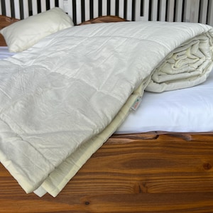 May include: A white quilted comforter folded on a wooden bed frame. The comforter has a subtle texture and a light cream color.