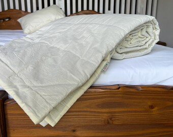 May include: A white quilted comforter folded on a wooden bed frame. The comforter has a subtle texture and a light cream color.