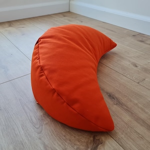 May include: A crescent moon-shaped pillow in a solid, vibrant orange fabric. The pillow features a smooth, matte texture and a curved design that tapers to pointed ends, with visible stitched seams along the outer edge of the curved form.