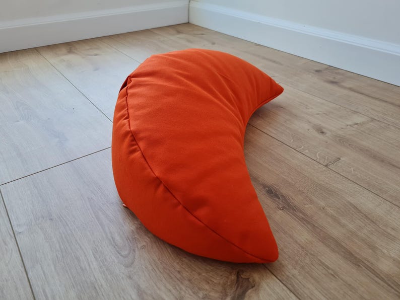 May include: A crescent moon-shaped pillow in a solid, vibrant orange fabric. The pillow features a smooth, matte texture and a curved design that tapers to pointed ends, with visible stitched seams along the outer edge of the curved form.