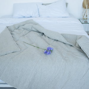 May include: A gray linen duvet cover with a single purple flower on top. The duvet is folded back on a white bed with white pillows.
