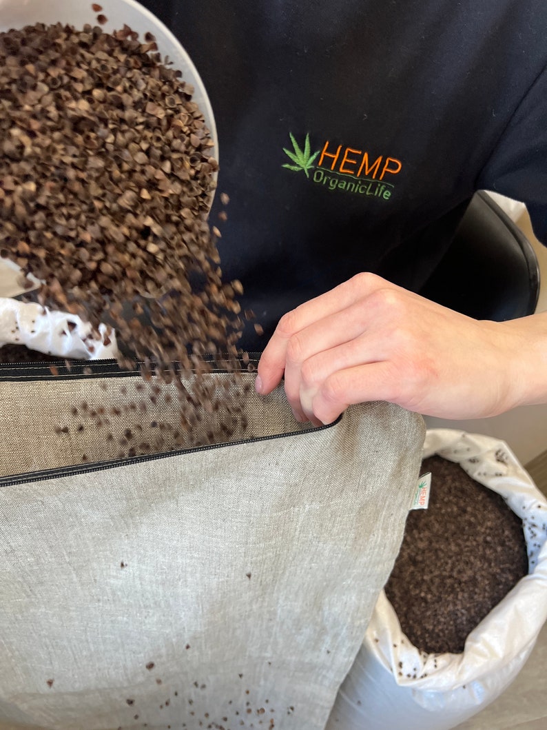 May include: A person is pouring buckwheat hulls into a large, light-colored canvas bag. The bag is sitting on a table and has a black zipper. The person is wearing a black shirt with a green hemp leaf logo that says "HEMP OrganicLife".