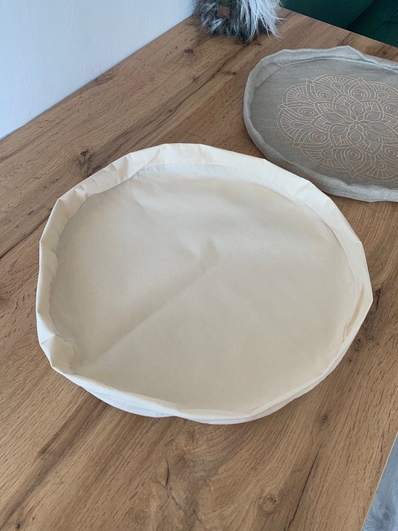 May include: A round, off-white canvas tray with a slightly raised edge. The tray is empty and sitting on a wooden surface.