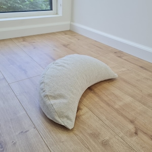 May include: A crescent-shaped meditation cushion made of light beige fabric. The cushion is sitting on a light wood floor.