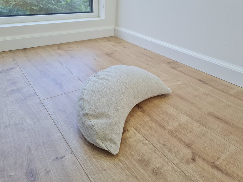 May include: A crescent-shaped meditation cushion made of light beige fabric. The cushion is sitting on a light wood floor.