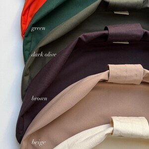 May include: A collection of fabric bags stacked vertically, featuring a range of colors labeled as dark blue, orange, green, dark olive, brown, beige, and natural. Each bag has a visible top handle strap and a smooth, solid-colored textile finish.