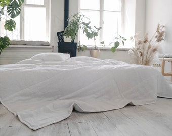 May include: A white linen duvet cover on a bed with white pillows. The bed is in a room with white walls and large windows. There are plants in the room, including a monstera plant in a pot on the floor.