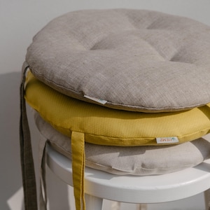 May include: A stack of three round seat cushions featuring a tufted design. The top cushion is beige, the middle is mustard yellow, and the bottom is light gray. Each cushion includes fabric ties on the sides and a small white brand label.