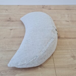 May include: A crescent-shaped meditation cushion made of light gray fabric. The cushion is on a light wood floor.