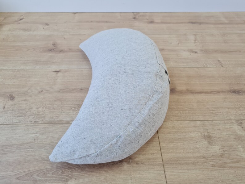 May include: A crescent-shaped meditation cushion made of light gray fabric. The cushion is on a light wood floor.