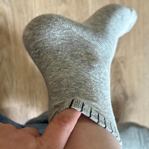 May include: Close-up of a grey sock on a foot. The sock is made of a ribbed knit fabric and has a folded cuff with black stripes. A finger touches the cuff of the sock.