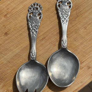 May include: Two silver-toned metal serving utensils, a salad fork and a spoon, featuring ornate handles with a relief design of a sailing ship and intricate scrollwork. The handles are topped with a decorative crown-like motif.