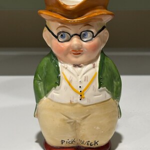 May include: A ceramic character jug shaped like a person wearing a brown top hat, round black glasses, a green coat, a white vest with a yellow chain, and tan trousers. The base of the figure features the black text Pick Wick printed on the front.