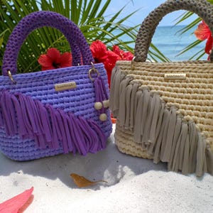 May include: Two handmade crochet handbags. One is lavender with matching fringe and a braided handle. The other is beige with matching fringe and a braided handle. Both have gold-coloured hardware and the word "handmade" on a small tag.