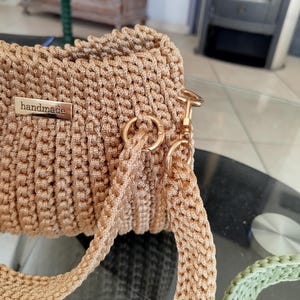 Puede incluir: Bolso beige hecho a mano con ganchillo, de forma rectangular. El bolso presenta una textura tejida, un cierre met&aacute;lico dorado y una correa a juego. Una peque&ntilde;a etiqueta met&aacute;lica dice "handmade".