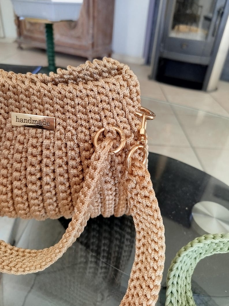 Puede incluir: Bolso beige hecho a mano con ganchillo, de forma rectangular. El bolso presenta una textura tejida, un cierre met&aacute;lico dorado y una correa a juego. Una peque&ntilde;a etiqueta met&aacute;lica dice "handmade".