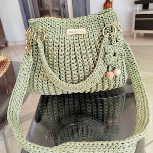 Puede incluir: Bolso de mano de ganchillo hecho a mano, de color verde salvia. El bolso tiene forma rectangular, herrajes dorados, una correa trenzada y un peque&ntilde;o adorno floral. La palabra "handmade" se muestra en una peque&ntilde;a placa dorada.
