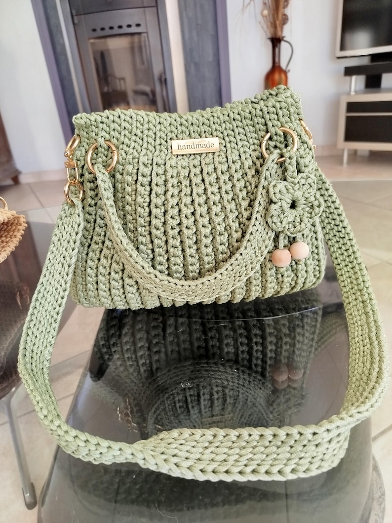 Puede incluir: Bolso de mano de ganchillo hecho a mano, de color verde salvia. El bolso tiene forma rectangular, herrajes dorados, una correa trenzada y un peque&ntilde;o adorno floral. La palabra "handmade" se muestra en una peque&ntilde;a placa dorada.
