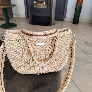 Op de afbeelding: Beige gehaakte handtas met een bruine bies en een lange, gevlochten riem. De tas heeft een rechthoekige vorm met franjes aan de voorkant. Een klein label met het woord "handmade" is zichtbaar.