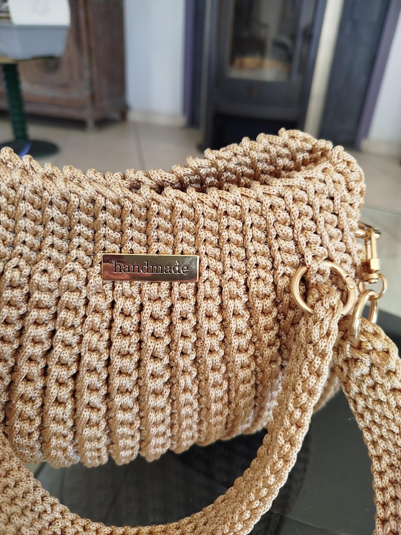 Puede incluir: Bolso beige hecho a mano con ganchillo, de forma rectangular. El bolso presenta una textura tejida y una etiqueta met&aacute;lica dorada con la inscripci&oacute;n "handmade". Tiene una correa a juego y herrajes dorados.